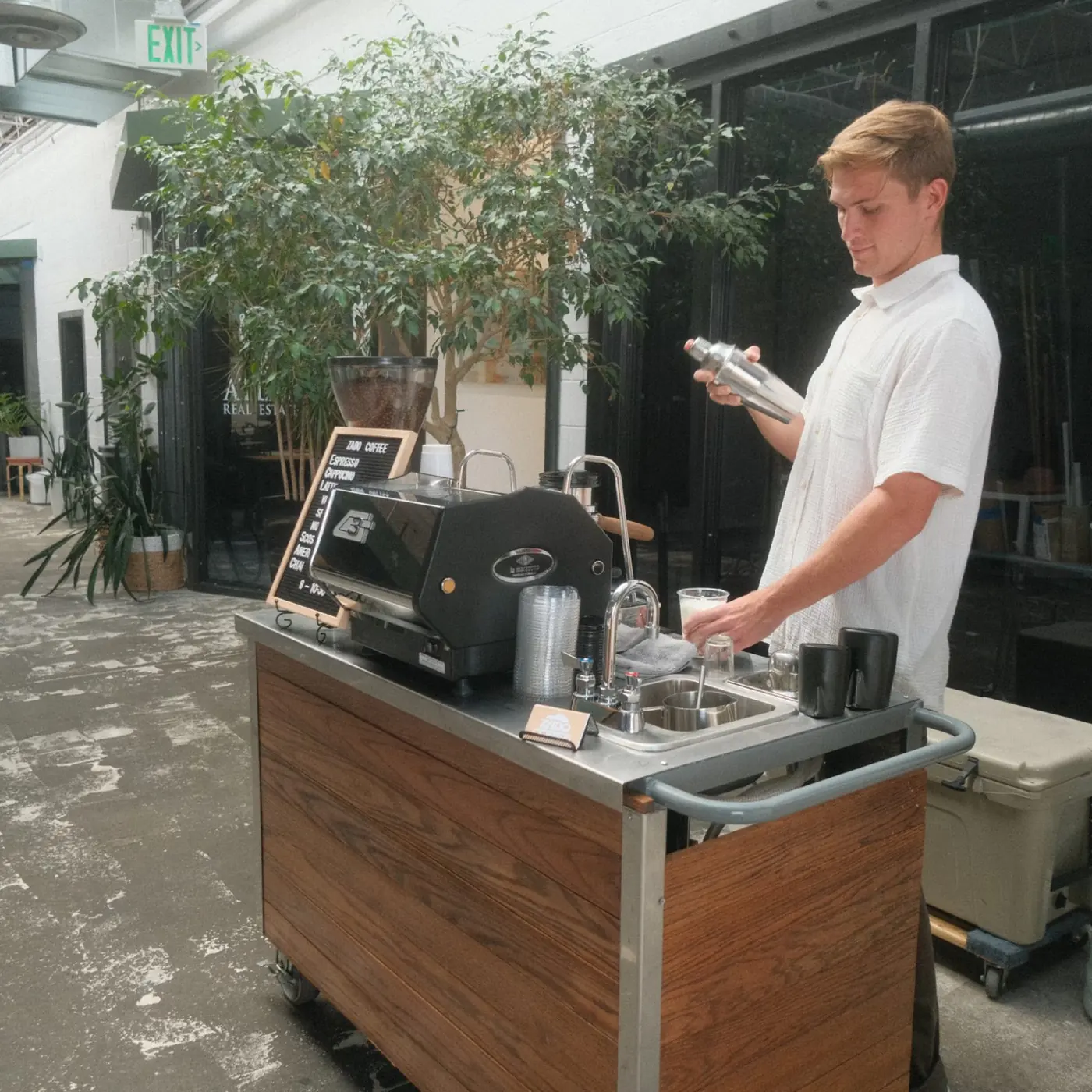 A Zado barista working at the wooden cart outside, with a big tree behind